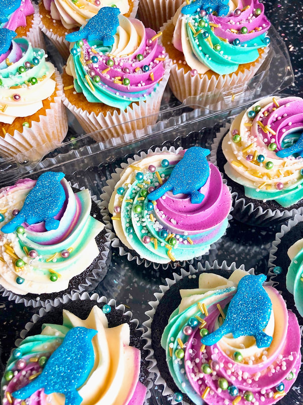 Cupcake Menu Photo: chocolate cupcakes topped with a swirl of white, teal, and purple frosting, metallic pink, gold, and teal sprinkles, and blue glittery fondant birds