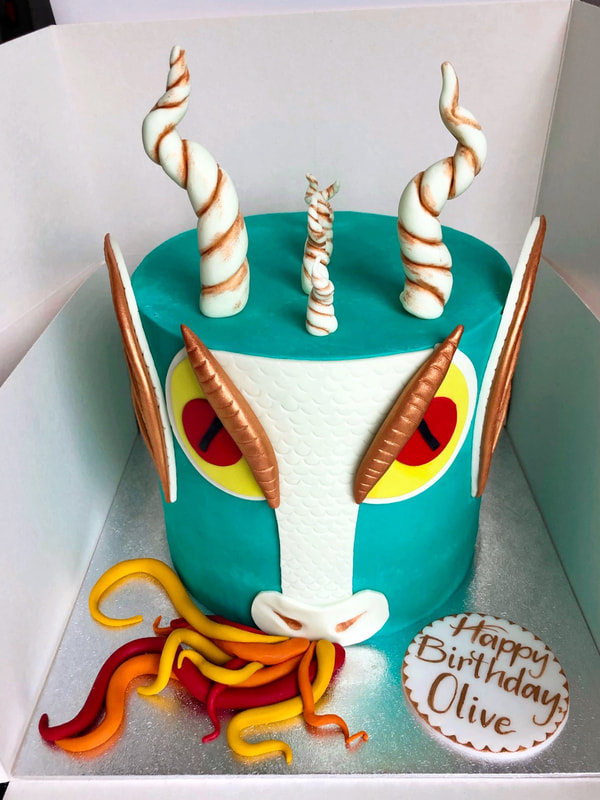 CAKE MENU: pictured is a custom dragon cake. It is teal with light green and bronze horns, wings, and eyebrows. It has big neon yellow and red eyes, and a light green snout with red, orange, and yellow flames coming out of its mouth.